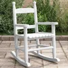 A white wooden rocking chair for children is placed on a paved patio in front of lush green bushes. Náhľad obrázku produktu