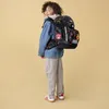 A young boy wearing a denim jacket and light-colored pants stands with his back to the camera, wearing a large, colorful backpack. The background is a plain, light yellow. Anteprima dell’immagine prodotto