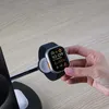 A hand is holding an Apple Watch over a charging stand. Produktbild-Vorschau