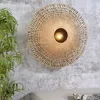 A woven wall lamp with a light bulb is mounted on a textured gray wall, with a green plant in the foreground and a sofa visible at the bottom. Anteprima dell’immagine prodotto