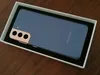 A smartphone is placed inside a white box, which is on a wooden surface. Produktbild-Vorschau