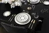 A formal dining table setting with black tablecloth, white and gold patterned plates, gold cutlery, and a floral arrangement. Náhled obrázku produktu