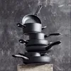 A stack of black pots and pans against a textured dark gray background. Náhled obrázku produktu