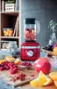 A red KitchenAid blender is shown on a countertop with various fruits and jars, suggesting a healthy lifestyle or recipe preparation. Produktbild-Vorschau