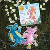 A children's game box and two dragon figures are laid out on grass with daisies. Produktbild-Vorschau