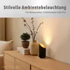 A black table lamp with a golden interior is placed on a wooden cabinet next to a vase with branches and a small potted plant. Produktbild-Vorschau