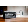 A black digital radio is placed on a wooden table in front of a blurred kitchen background with a faucet. Produktbild-Vorschau