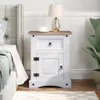 A white bedside table with a wooden top, a drawer, and a cabinet door, placed next to a bed. A lamp and a cup are on top of the table. Produktbild-Vorschau