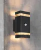 A black outdoor wall light with gold accents is mounted on a textured gray concrete wall. Produktbild-Vorschau