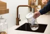 A person is filling a water filter pitcher under a copper faucet in a modern kitchen. Produktbild-Vorschau