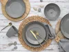 A table setting with grey stoneware plates, a mug, cutlery, and a napkin ring, arranged on a wooden surface with decorative stones. Náhľad obrázku produktu