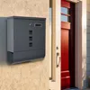 A dark gray metal mailbox is mounted on a textured beige wall next to a red door. Produktbild-Vorschau