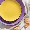 A close-up overhead view of two colorful ceramic plates, one yellow and one purple, resting on a textured white cloth. Several cookies are scattered on the cloth next to the plates. Produktbild-Vorschau