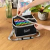 A person is opening a bag of colorful markers on a wooden table, with a plant and a drawing in the background. Náhľad obrázku produktu