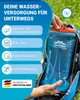 A person's hand is holding a blue water bladder with a drinking tube, which is partially inserted into a blue backpack. The background shows greenery. Produktbild-Vorschau
