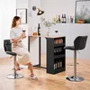 A woman sits at a bar table, holding a glass of wine, with bar stools and bottles of wine visible. Produktbild-Vorschau