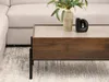 A close-up shot of a modern coffee table with a wooden base and a beige top, placed on a patterned rug next to a sofa. Decorative items, including a vase with greenery and a black abstract sculpture, are on the table. Náhled obrázku produktu
