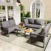 A patio set with gray cushions and dark frames is arranged on a light-colored rug on a tiled patio, with a white arched doorway and greenery in the background. Produktbild-Vorschau