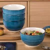 A stack of blue ceramic bowls and one bowl filled with salad are displayed on a wooden surface, with a blurred background of wooden cabinets. Náhled obrázku produktu