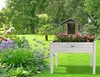 A raised garden bed filled with pink flowers sits in a lush green garden with a small wooden shed in the background. Voir les images du produit