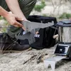 A person is holding a metal grill plate over a portable stove in an outdoor setting. Produktbild-Vorschau