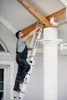 A man is using a ladder to reach a wooden beam on the exterior of a building. Produktbild-Vorschau