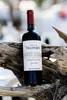 A bottle of red wine is placed on a weathered piece of driftwood, with a blurred background of trees and sky. Produktbild-Vorschau
