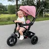 A baby sitting in a pink tricycle on a paved path outdoors. Produktbild-Vorschau