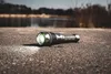 A flashlight lies on a textured surface with a body of water and trees in the background. Produktbild-Vorschau
