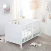 A white baby crib in a nursery room with artwork on the wall and a window in the background. Produktbild-Vorschau