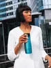 A woman holding a turquoise travel mug in front of a modern building. Produktbild-Vorschau