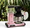 A pink Moccamaster coffee maker is shown on a white surface with lush green plants in the background. Náhľad obrázku produktu