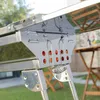 A close-up view of a portable barbecue grill, showing its metallic structure and some of its components. The background is blurred, showing a garden setting with tables and chairs. Produktbild-Vorschau
