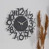 A modern black wall clock with a unique design is mounted on a light gray wall, with dried grass stalks to the right. Produktbild-Vorschau