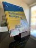 A cookbook is displayed on a wooden stand on a reflective surface, with a blurred background suggesting an indoor setting. Produktbild-Vorschau