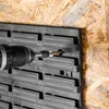 A close-up shot of a black plastic tool organizer panel being attached to a wooden wall with a screw and a drill. Produktbild-Vorschau