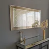 A rectangular mirror with a decorative frame hangs on a gray wall above a mirrored console table. Decorative items, including an hourglass and a vase with dried flowers, are placed on the table. Produktbild-Vorschau