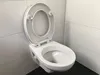 A white toilet with the lid up, set against a tiled wall and dark floor. Produktbild-Vorschau