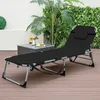 A black folding lounge chair is set up on a wooden deck, with a small table and drinks in the background, surrounded by plants. Podgląd zdjęcia produktu