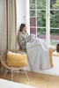 A woman sits by a window, wrapped in a blanket, looking out at the trees. A yellow pillow rests on a chair next to her. Produktbild-Vorschau