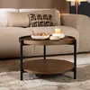 A round, two-tiered coffee table with a dark oak finish and black metal legs, placed on a rug in a living room setting.