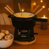 A fondue set with cheese, forks, and a burner on a wooden table, with fairy lights and a candle in the background. Podgląd zdjęcia produktu