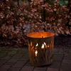 A rusty metal fire basket with flame cutouts is lit with flames, sitting on a paved patio in front of a blurred background of brown leaves. Produktbild-Vorschau