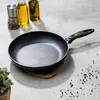 A frying pan is placed on a concrete surface with cooking ingredients and bottles of oil in the background. Produktbild-Vorschau