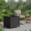 A dark gray wooden planter box sits on a patio, surrounded by rocks and lush green plants. A chair and small tables are visible in the background. Náhľad obrázku produktu