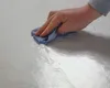 A hand is wiping a white surface with a blue cloth, leaving wet streaks. Produktbild-Vorschau