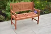A wooden garden bench is placed on a paved patio in front of a garden with plants and flowers. Produktbild-Vorschau