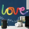 A neon LED sign spelling 'LOVE' in rainbow colors is mounted on a dark blue wall above a desk. On the desk, a black portable speaker is placed next to a potted plant, a clock, and some pencils. Musical notes are drawn on the wall behind the speaker. Anteprima dell’immagine prodotto