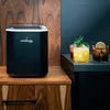 An ice maker is placed on a wooden cabinet next to three cocktails on a countertop. Náhled obrázku produktu