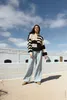 A woman stands outdoors in front of a white building with a blue sky and clouds above. Produktbild-Vorschau